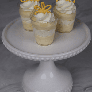 Three small CO-104 Mango Shooters of layered yellow and white mousse, topped with whipped cream and yellow chocolate decorations, displayed on a white beaded-edge cake stand against a marble background.