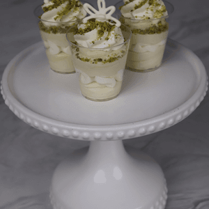 Three CO-103 Pistachio Shooters, small dessert cups of creamy white mousse topped with crushed pistachios and white chocolate decor, are displayed on a cake stand against a marble background.