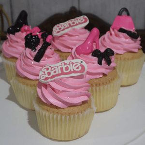 Six CC-030B Gold cupcakes with chocolate filling, topped with decorative plastic items such as a Barbie logo, high heel shoe, purse, bow, and lipstick, are arranged on a white plate.