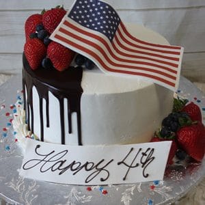 JC-000: A round cake with white frosting and chocolate drip, topped with strawberries, blueberries, and an American flag. "Happy 4th" is written on a white plaque. The cake sits on a silver platter with star sprinkles.
