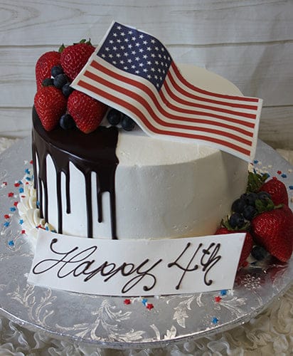 JC-000: A round cake with white frosting and chocolate drip, topped with strawberries, blueberries, and an American flag. "Happy 4th" is written on a white plaque. The cake sits on a silver platter with star sprinkles.