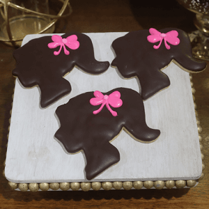 Three CE-001g birthday cookies, shaped like a girl's head in profile with a ponytail, chocolate icing, and a pink bow, are displayed on a square wooden tray.