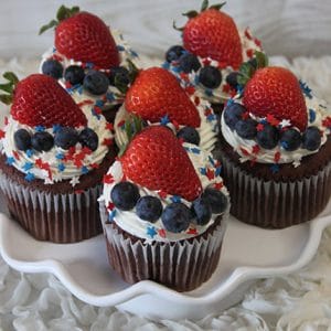 JC-025 Chocolate cupcakes with white chocolate mousse filling, topped with strawberries, blueberries, and red, white, and blue star-shaped sprinkles, are arranged on a white cake stand against a light background.