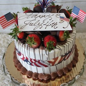 The JC-011 Strawberry Grand Marnier is a festive cake topped with chocolate strawberries, American flags, and a “Happy July 4th” topper, featuring red, white & blue accents plus piped chocolate frosting on a glass plate.