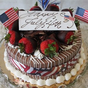 The JC-004 Chocolate/Oreo cake features chocolate-covered strawberries, chocolate shavings, and U.S. flags, topped with a "Happy July 4th" sign and festive decorations, and wrapped in a red, white, and blue ribbon.