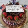 The JC-003 Chocolate/Chocolate cake is topped with chocolate-dipped strawberries, two mini American flags, a festive red, white, and blue streamer topper, and a white plaque reading "Happy July 4th!" in cursive.