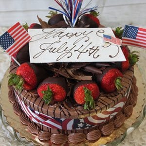 The JC-003 Chocolate/Chocolate cake is topped with chocolate-dipped strawberries, two mini American flags, a festive red, white, and blue streamer topper, and a white plaque reading "Happy July 4th!" in cursive.