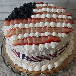 The JC-001 is a round cake on a glass platter, decorated to look like the American flag with blueberries for the blue field, sliced strawberries for red stripes, and piped white frosting for the white stripes.