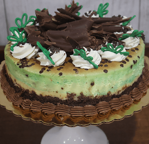 The GD-016 Mint Chocolate Chip Cheesecake features round green and brown layers, topped with whipped cream, chocolate shavings, green icing accents, and chocolate chips on a gold cake board and white stand.