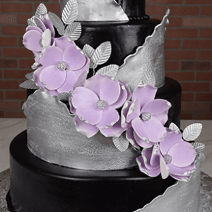 The WD-006 is a four-tier black cake featuring metallic silver bands swirling up each layer, with clusters of light purple flowers and silver leaves, set against a brick wall backdrop.