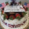 A festive JC-007 Pina Colada cake adorned with strawberries, chocolate, small American flags, red, white, and blue ribbon, plus a 'Happy July 4th' card in cursive sits on a glass platter.