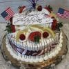 The JC-008 Lemon and Raspberry cake features festive strawberries, cream, lemon slices, chocolate drizzle, small U.S. flags, and a "Happy 4th of July!" sign on top, all presented elegantly on a glass platter.