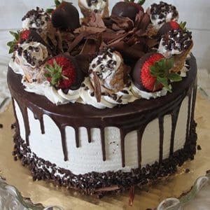 The BD-018E Cannoli Cake is a round cake with white frosting, chocolate drip, chocolate shavings, cannoli, whipped cream, and chocolate-covered strawberries, served on a glass platter.