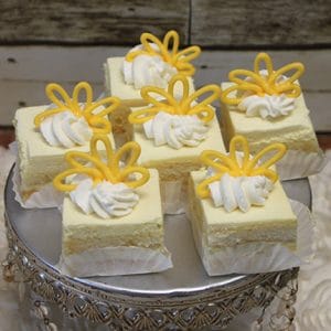 Six square Mango squares topped with whipped cream and yellow icing are arranged on a silver cake stand with crystal accents, each served in a white paper cup.