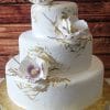 WD-016: Three-tiered white cake with gold leaf accents and large white sugar flowers with gold centers, displayed on a gold cake board against a wooden background.
