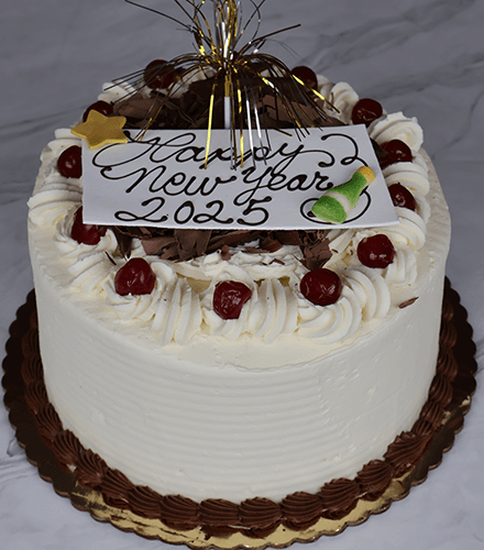The NY-004 Black Forest cake is a round white frosted treat adorned with whipped cream swirls, cherries, chocolate pieces, and a gold topper. A card atop the cake says “Happy New Year 2025” in chocolate icing.
