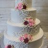 Product WC-009 is a three-tiered white wedding cake adorned with ornate icing swirls and clusters of pink and burgundy roses cascading down the side, presented against a light wooden background.
