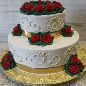 WC-010 is a two-tiered white cake adorned with gold ribbon, ornate icing swirls, and clusters of red frosting roses with green leaves, presented on a gold-patterned cake board.