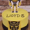 The CB-006I is a yellow cake featuring a black Transformers logo, a Bumblebee character topper, and "LIOYD 5" written in black icing—ideal for a child's fifth birthday. The cake is presented on a gold tray.