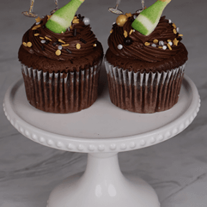 Two NY-018 Chocolate cupcakes with white filling, gold and white sprinkles, chocolate frosting, champagne bottle candies, and glittery champagne glass toppers, displayed on a white cake stand.