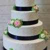 Product WC-023: A four-tiered white wedding cake with black ribbon bands, pink and white icing roses, green leaves, and dotted icing details, displayed against a light wooden background.