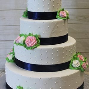 Product WC-023: A four-tiered white wedding cake with black ribbon bands, pink and white icing roses, green leaves, and dotted icing details, displayed against a light wooden background.