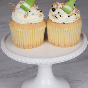 A NY-017 Gold cupcake with chocolate filling sits on a white cake stand, topped with white frosting, colorful sprinkles, and gold toppers shaped like sparkling champagne bottles and glasses.