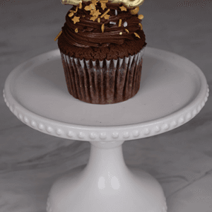 The NY-016 Chocolate cupcake with white filling, topped with gold star sprinkles and a gold 2025 topper, is displayed on a white decorative cake stand against a marble background.