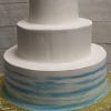 WC-014: A three-tiered white cake with smooth frosting; the bottom tier has blue watercolor-style stripes. Presented on a gold cake board, set against a light wooden background.