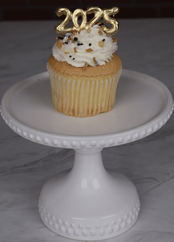 A NY-015 Gold cupcake with chocolate filling and gold sprinkles sits on a white cake stand, topped with a gold 2023 decoration.