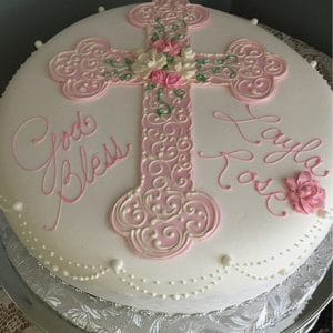 RC-047: Round white cake featuring a large ornate pink cross, white and pink flowers, and "God Bless Layla Rose" in pink icing, presented on a silver foil-covered board.