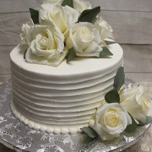 The BR-001 cake features white frosted, textured sides and is adorned with fresh white roses and green leaves, presented on a silver patterned cake board.