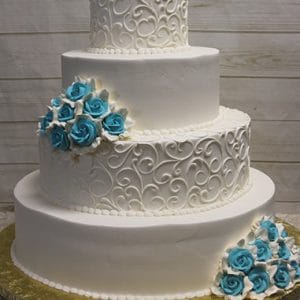 The WC-013 is a four-tiered white wedding cake featuring elegant swirl patterns and clusters of blue and white roses, displayed on a gold cake board against a light wooden background.