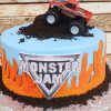 CB-010H is a round cake with a blue background, featuring orange flames, the Monster Jam logo, crumbled cookie “dirt,” and topped with a toy monster truck. It’s displayed on a silver base.