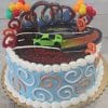 The SC-084 Hot Wheels is a round birthday cake with blue and white icing, orange swirls, a toy green car topper, looping racetrack decoration, balloon clusters, and red "Happy Birthday" text.