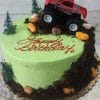 A green birthday cake topped with a red SC-085 Monster Truck toy, small trees, candy rocks, and chocolate crumbs as dirt. "Happy Birthday" is written in red icing on top.
