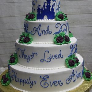 WC-019 is a four-tiered white wedding cake with purple and green flowers, blue icing that says “And they lived happily ever after,” and a blue castle silhouette on top.