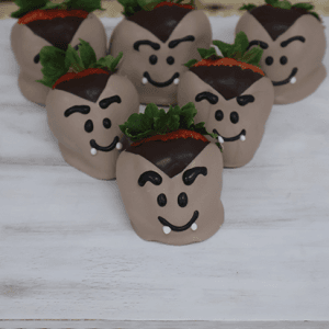 Six HW-021 strawberries are dipped in chocolate and decorated with cartoonish eyes, fangs, and expressive eyebrows to resemble vampires. They are arranged on a white surface for a festive presentation.