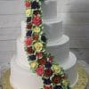 WC-020: Four-tiered white cake with cascading red, yellow, purple, and burgundy buttercream roses, displayed on a gold cake board against a wood background.