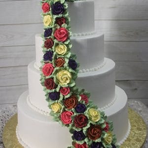 WC-020: Four-tiered white cake with cascading red, yellow, purple, and burgundy buttercream roses, displayed on a gold cake board against a wood background.