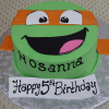 A green CB-005G birthday cake shaped like a Teenage Mutant Ninja Turtle face with an orange mask, smiling mouth, and "Hosanna" written on it. A front sign says "Happy 5th Birthday.