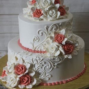 The WC-018 is a two-tiered white wedding cake with intricate swirl designs, topped with white and pink fondant roses clusters, set on a gold cake board.