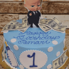 The CB-010I is a blue birthday cake with white polka dots, a large number one, dollar bills, a baby bottle, Boss Baby topper, and “Happy Birthday Tamari” written in blue icing.