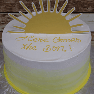 The BB-076 cake features white and yellow frosting, a sun design, and “Here Comes the Son!” written in yellow icing. It’s displayed on a lightly patterned surface with a rustic wooden background.