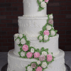 WC-000L is a four-tiered white wedding cake adorned with detailed piping and cascading pink, white, and light purple buttercream roses with green leaves, beautifully displayed against a dark brick backdrop.