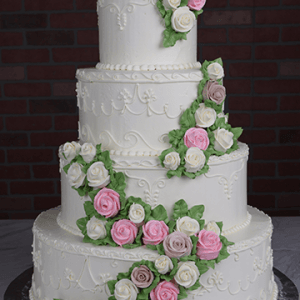 WC-000L is a four-tiered white wedding cake adorned with detailed piping and cascading pink, white, and light purple buttercream roses with green leaves, beautifully displayed against a dark brick backdrop.
