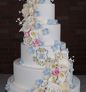 The WD-000E is a tall, five-tiered white wedding cake with cascading white, pink, and blue sugar flowers and a floral crest on the middle tier, displayed against a dark brick wall background.
