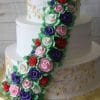 WC-016 is a three-tiered white cake featuring gold vine accents and a cascade of colorful buttercream roses in red, purple, pink, and white, finished with green leaves.