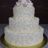 The WC-000B is a three-tiered white wedding cake with ornate swirl patterns, topped with light pink and white roses, and presented on a gold base against a brick wall backdrop.