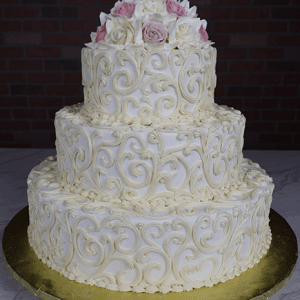 The WC-000B is a three-tiered white wedding cake with ornate swirl patterns, topped with light pink and white roses, and presented on a gold base against a brick wall backdrop.